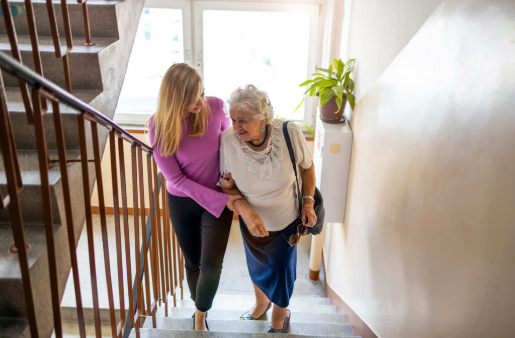 A young woman helping a senior woman walk up a stairway, holding her arm for assistance.