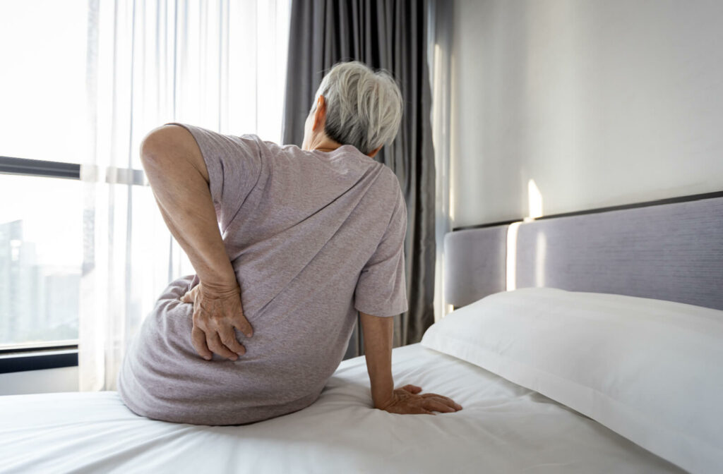 A senior woman suffering from back pain is sitting at the side of her bed having a hard time standing.