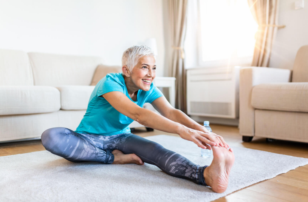A senior woman stretching to reach her toes on her left foot while sitting on a grey rug.