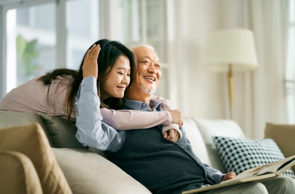 Older adult sitting on a couch smiling while being hugged by a loved one in a bright and cozy living room