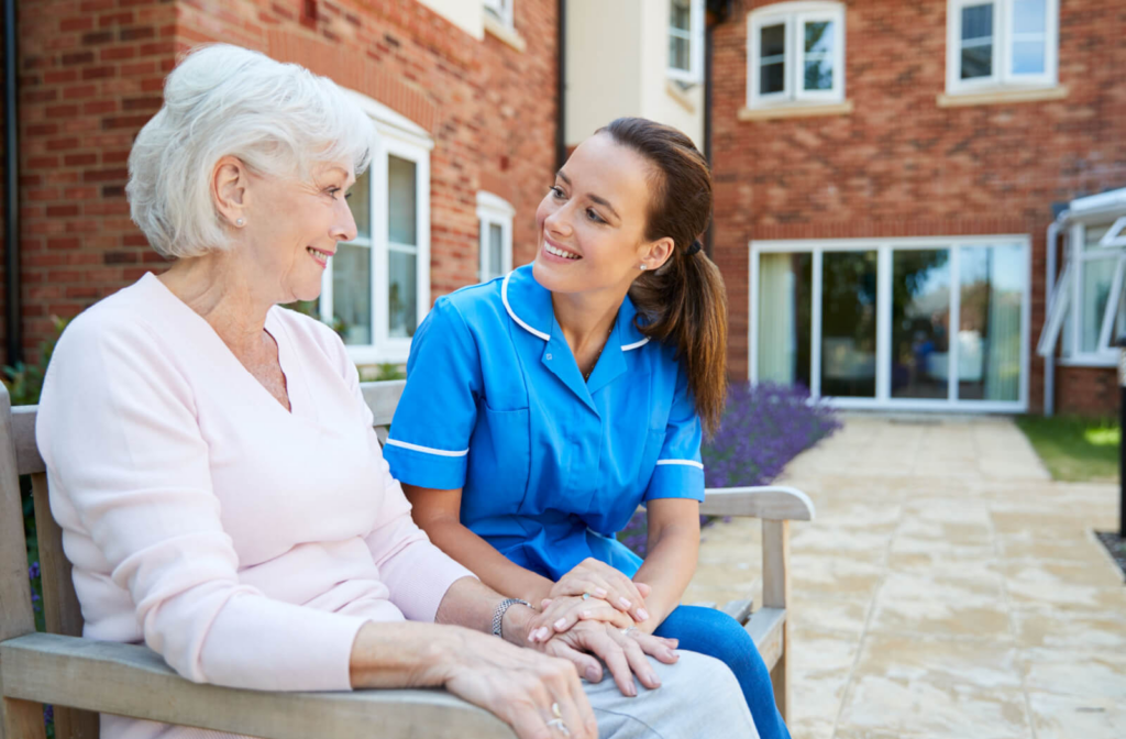 An older adult smiles while sitting on a bench in a courtyard in assisted living as a caregiver stops to sit and check in on them