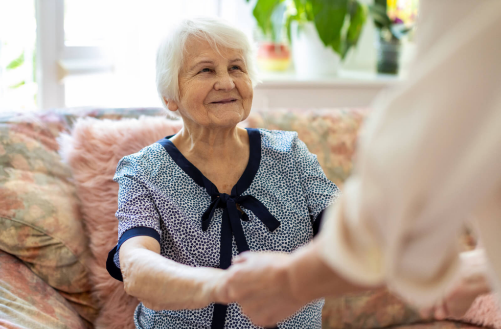 An out-of-frame caregiver helps an appreciative older resident stand up from a comfortable couch in their living room in assisted living