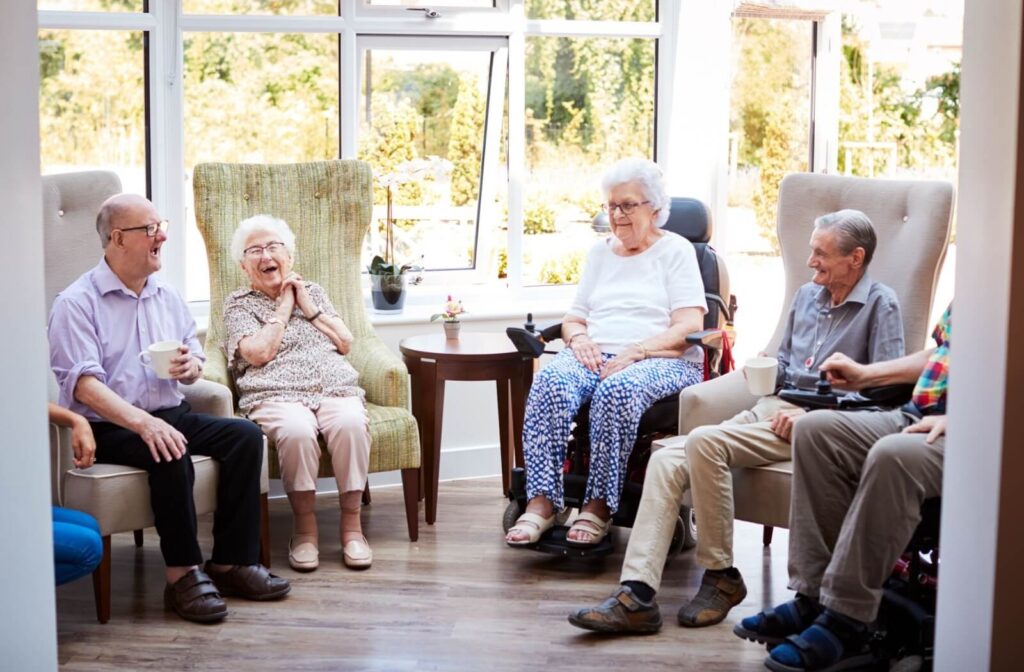 Residents in senior living sit in a semi-circle in comfortable chairs in a lounge in front of a beautiful sunlit window